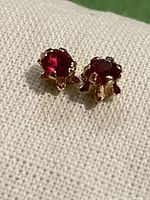 Front view of two small stud earrings with red stones set in yellow gold claw settings placed on a white fabric background.