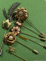 Eight assorted vintage hat pins arranged on green background showing various floral, cameo, and leaf designs with pearl and purple accents
