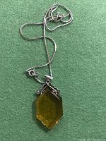 Photo showing entire pendant hanging on the chain on green fabric background, displaying geometric amber-colored stone and gold metal setting.