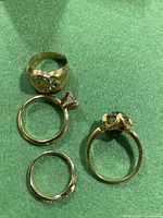 Four assorted vintage gold tone rings displayed on green fabric, showing front and side views emphasizing stone settings and band details.