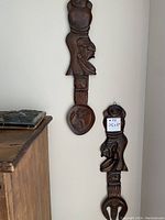 Pair of carved wood fork and spoon wall hangings displaying front carving details, hung on a beige wall.