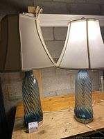Photo of both table lamps side by side on a wooden surface against a cinder block wall, showing the full lamps with shades on.