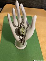 Photo of the pocket watch displayed on a white ceramic hand with chain draped over fingers to show length and style.