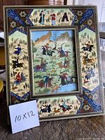 Full view of framed painting with intricate decorative painted frame, showing hunting scenes and floral patterns, with visible damage on top.