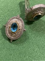 Close-up of one oval clip-on earring with blue faceted stone and detailed silver and gold engraved setting.