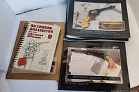 Four firearms-related books displayed together on a white surface: one spiral-bound ballistics book and three firearms auction catalogs with vintage revolver images.