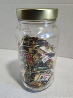 Glass jar showing front view of assorted pins inside with diverse designs, including flags and symbols.