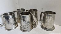Five pewter mugs shown together displaying etched designs including a police badge and a map.