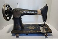 Side view of vintage Singer sewing machine showing black paint with gold floral decorations and Singer logo on arm. Manual handwheel on side.