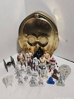 Front view of Star Wars toys and figurines arranged in rows in front of the gold C-3PO shaped metal tin including characters, vehicles and game pieces.