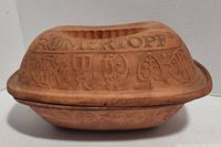 Side view of terracotta casserole showing embossed ROMERTOPF and decorative symbols on lid and base.