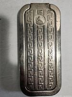 Close-up of the metal case showing the geometric Greek key-style patterns and the circular medallion with a seated figure and 'ROLLS' branding.