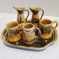 Top side view of five ceramic collectible pieces on a decorative tin tray, showing vintage car images and landmark designs.