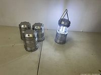 Four silver camping lanterns displayed together, one lantern turned on showing bright LED light, with collapsible hanging handles.