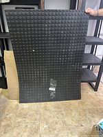 Full view of rectangular black rubber shop mat with circular textured grips held vertically by a person showing size and texture.