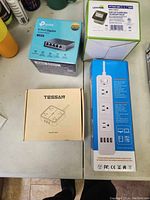 Photo of four boxed electronic items including the TP-Link 5-port Gigabit switch, Leviton pop-up floor box RT receptacle, Tessan power adapter plug, and a white power bar with USB ports.