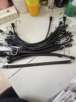 Photo showing a pile of multiple black rubber tarp straps with silver metal hooks on both ends arranged on a white surface to display quantity and design.