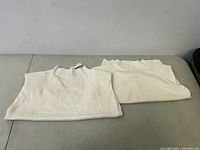 Two white ribbed tops folded on a table, showing fabric texture and high neckline.