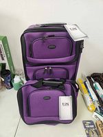 Two purple luggage pieces, a larger suitcase and a smaller carry-on bag, both with front zip pockets and US Traveler tags.