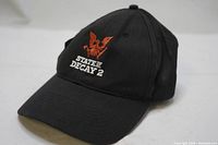 Front view of the black baseball cap displaying the embroidered orange eagle logo and white 'State of Decay 2' text on front panel.