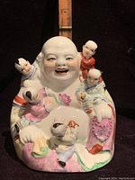 Front view of the porcelain figurine showing the Laughing Buddha smiling with five children climbing around him, colored in bright traditional robes.