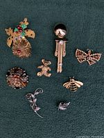 Full set of 8 assorted brooches and pins arranged on a dark fabric background showing various designs including butterfly, flower, bee, teddy bear, and abstract shapes.