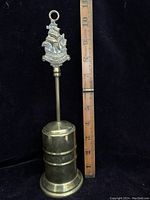 Front view of vintage brass retractable fireplace brush with ship emblem and ruler for scale.