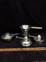Front view of all four warmers/burners including Kinox stainless piece with wood handle pan, metal chafing dish warmer, and two silver plated wick warmers, with ruler for scale.