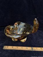 Photo of amber mid-century modern art glass bowl showing flowing freeform shape with curled ends and amber/gold coloration. Ruler for size reference approximately 7-8 inches long.