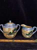 Photo showing both covered sugar bowl and creamer together with a ruler for size reference against black background.