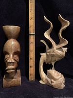 Photo showing two wooden carvings with a measuring ruler behind for scale. The carving on the left is a tribal bust, about 7 inches tall, with a bowl-shaped top and prominent facial features. The carving on the right is a Bali style kissing couple with abstract upward flowing shapes, about 8 inches tall.