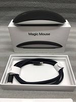 Apple Magic Mouse on top of original box showing curved design and Apple logo, with braided charging cable inside box.