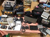 Wide shot showing large assortment of untested electronics including cameras, camcorders, headphones, remote controls, and lighting on a table.