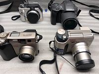 Four cameras on a flat surface including silver Panasonic Lumix DMC-LC40, Olympus and Canon cameras