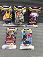 Five sealed Pokémon Trading Card Game booster packs arranged in two rows on gray fabric background. Packs include Silver Tempest (two packs), Brilliant Stars (one pack), and Lost Origin (two packs).