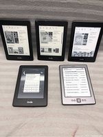 Five Kindle e-readers arranged on a gray fabric surface, various screen displays visible, including library and lock screen states.