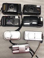 Seven vintage compact cameras arranged on a surface showing their fronts and varying brands and models.