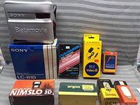 Photo of Sony Betamovie camcorder case and various camera accessory boxes including Sony LC-810 tape box, Argus camera film box, batteries, and flash packaging.