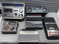 Top view of seven vintage portable electronics on a grey surface showing cassette players, radios, and a clock radio