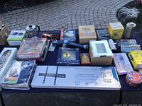 Photo of all items arranged on a black cloth with pavement background including kitchenware, electronics, fabrics, boxed products, cables, clock, and TV mount.