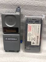 Front view of vintage Motorola flip phone with antenna and small screen, beside extra battery pack with Motorola label.