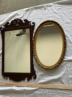 Two decorative wall mirrors laid flat on white sheet, side by side, larger rectangular wooden mirror with carved floral detail on left, smaller oval framed mirror with floral gold frame on right.