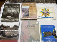 Five books arranged on ground showing front covers: 'The Dust Bowl,' 'Where the Old West Stayed Young,' 'The Dust Bowl Through the Lens,' 'Colorado,' and 'Windmills of the West.'