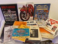 Photo showing the assortment of Harley-Davidson related books, manuals, and memorabilia on a table, including hardcover and paperback books, repair guides, and manuals.