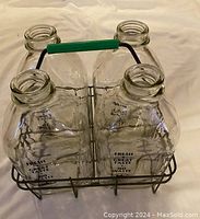 Four clear glass milk bottles embossed with text placed inside a metal carrier with green handle, visible from an angle.