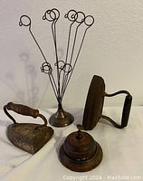 Photo showing two used cast iron sad irons, metal bell, and wire photo stand on a white surface against light wall.