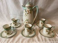 Full set photo showing pot with lid standing behind 5 cups placed on 5 saucers, floral decoration visible