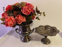 Front view of silver plate ice bucket filled with artificial pink roses and peonies, next to a decorative silver plate compote bowl