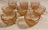 Eight pink/peach translucent glass cups with ornate embossed floral and swirl patterns, arranged on a light surface showing slight color variations and transparency.