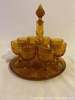 Front view of the assembled amber sandwich glass decanter with stopper, tray, and eight glasses arranged around the decanter on the tray.